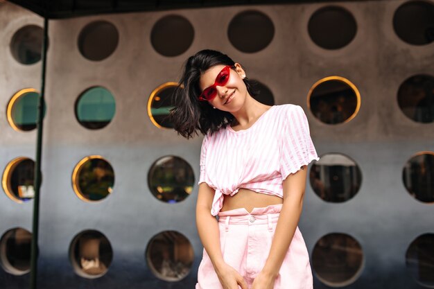 Felice donna alla moda che guarda l'obbiettivo su sfondo urbano. Colpo esterno di splendida donna in occhiali da sole rosa in posa con il sorriso.