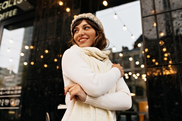 Felice donna amabile in abito invernale bianco che cammina per strada con un sorriso felice su sfondo con luci di Natale Foto di alta qualità