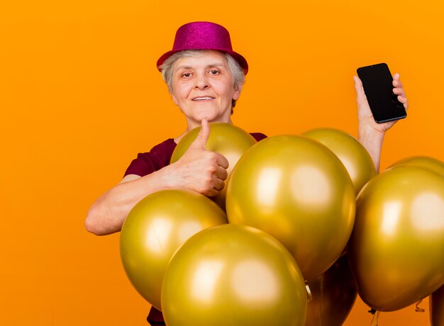 Felice donna anziana che indossa il cappello da festa sta con i pollici di palloncini di elio e tiene il telefono isolato sulla parete arancione