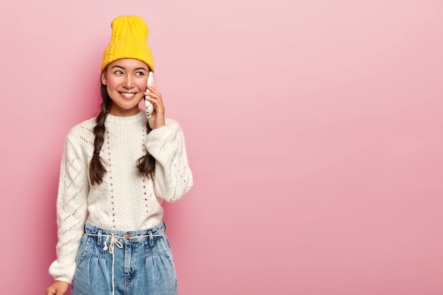 Felice donna asiatica parla tramite smartphone, ha una conversazione piacevole, chiama un amico, indossa un maglione bianco caldo e jeans, guarda da parte con un sorriso, posa su un muro rosa