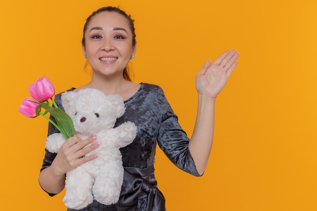 Felice donna asiatica tenendo il mazzo di tulipani rosa e orsacchiotto sorridendo allegramente celebrando la giornata internazionale della donna in piedi sopra la parete arancione