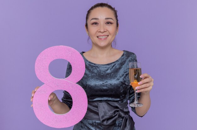 Felice donna asiatica tenendo il numero otto realizzato in cartone e un bicchiere di champagne guardando davanti sorridendo allegramente per celebrare la giornata internazionale della donna in piedi sopra la parete viola