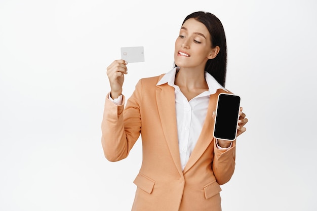 Felice donna aziendale guardando la sua carta di credito che mostra lo schermo dello smartphone in piedi in tuta su sfondo bianco