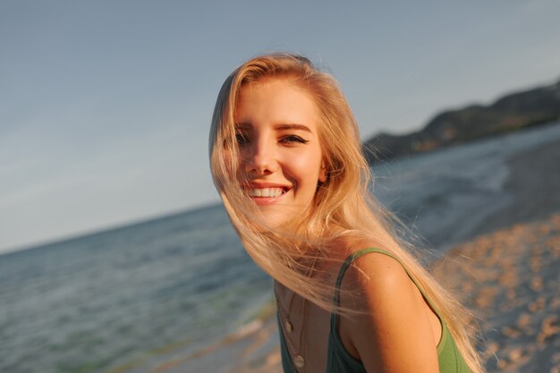 Felice donna bionda con un sorriso perfetto divertirsi sulla spiaggia assolata, guardando la fotocamera, correre e ballare