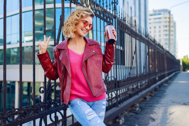 Felice donna bionda in posa su strade moderne, bere caffè o cappuccino. Elegante abito autunnale, giacca di pelle e maglione lavorato a maglia. Occhiali da sole rosa.