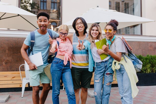 Felice donna bionda indossa jeans con fori in posa all'aperto vicino ad amici sorridenti. Ritratto all'aperto di studenti soddisfatti che tengono laptop e zaini in mattinata.