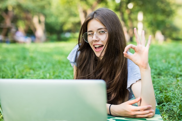 Felice donna bruna in occhiali sdraiato sull'erba nel parco con computer portatile e mostrando segno ok