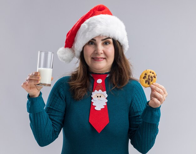 Felice donna caucasica adulta con cappello santa e cravatta santa tenendo un bicchiere di latte e cookie isolato su sfondo bianco con spazio di copia