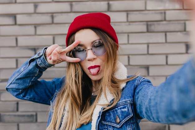 Felice donna caucasica in posa con la lingua fuori sul muro di mattoni. Ragazza attraente in cappello rosso che fa selfie con gli occhi chiusi.