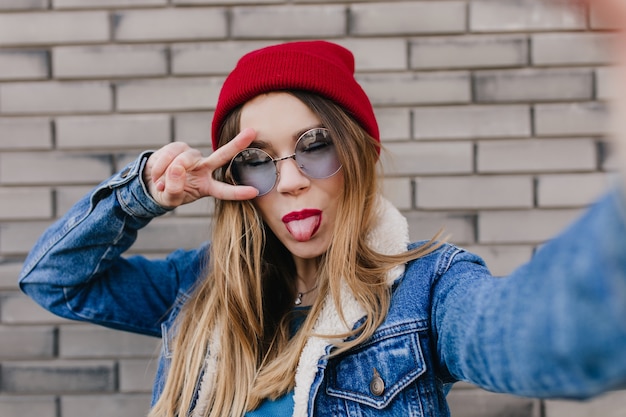 Felice donna caucasica in posa con la lingua fuori sul muro di mattoni. Ragazza attraente in cappello rosso che fa selfie con gli occhi chiusi.