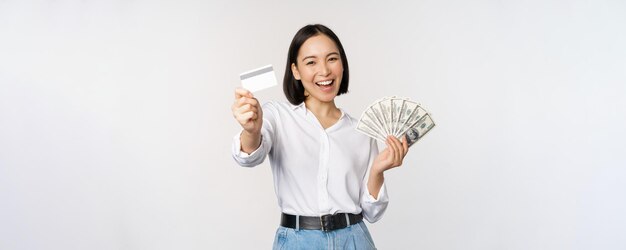 Felice donna coreana in possesso di carta di credito e denaro dollari sorridente e ridendo in posa su sfondo bianco per studio