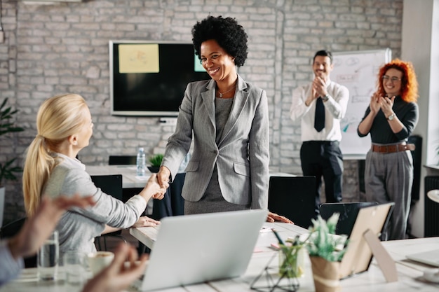 Felice donna d'affari afroamericana che stringe la mano alla sua collega in ufficio mentre altri colleghi li applaudono