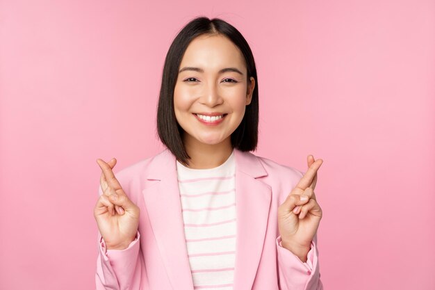 Felice donna d'affari asiatica incrociare le dita per buona fortuna che desiderano pregare e sperare di sorridere alla telecamera in piedi in tuta su sfondo rosa