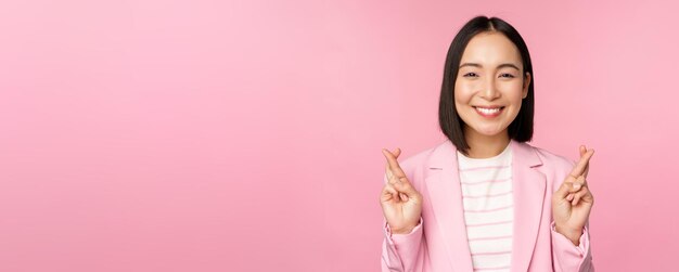 Felice donna d'affari asiatica incrociare le dita per buona fortuna che desiderano pregare e sperare di sorridere alla telecamera in piedi in tuta su sfondo rosa