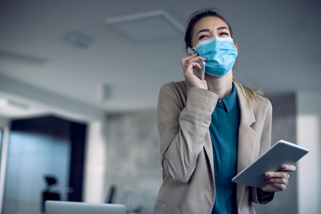 Felice donna d'affari con maschera facciale che parla al telefono mentre si utilizza il touchpad in ufficio