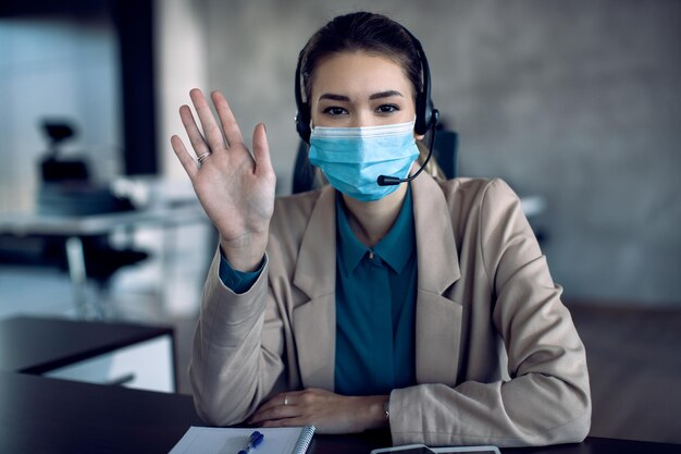 Felice donna d'affari con maschera facciale che sventola durante la teleconferenza in ufficio