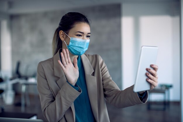 Felice donna d'affari con maschera facciale che sventola durante la videochiamata sul touchpad in ufficio