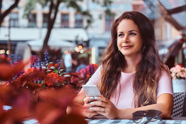 Felice donna d'affari di mezza età con lunghi capelli castani tiene uno smartphone mentre è seduto in un caffè all'aperto.