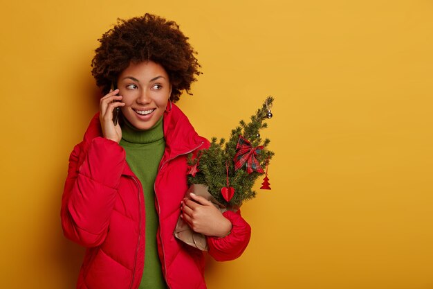 Felice donna dai capelli ricci parla al telefono, tiene un piccolo abete decorato per Natale