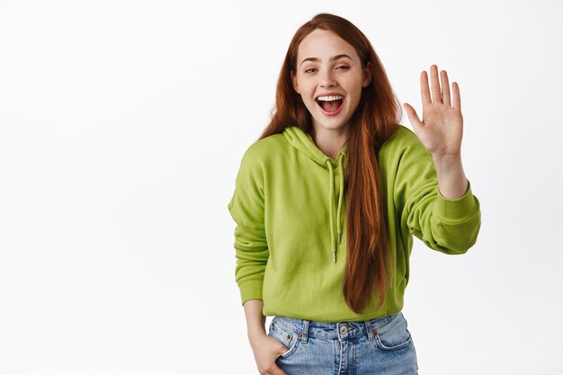 Felice donna dai capelli rossi saluta, agitando la mano alzata gesto di saluto, sorridendo amichevole, in piedi sul bianco