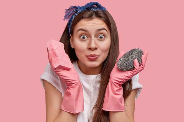 Felice donna dai capelli scuri si prende cura dei sanitari e dell'igiene, indossa guanti di gomma, tiene due mop in mano, si prepara a pulire il bagno sporco, indossa una maglietta bianca