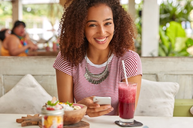 Felice donna dalla pelle scura con capelli croccanti, legge le notizie sul sito web, si connette a Internet wireless alla caffetteria, beve frullati freschi, posa al ristorante con terrazza, installa l'app, indossa una maglietta, una collana