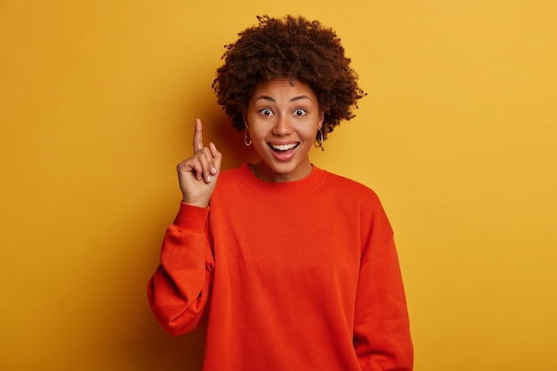 Felice donna dalla pelle scura con taglio di capelli afro consiglia un prodotto fantastico, indica soddisfatto, ti mostra un promo perfetto, vestito con un maglione rosso, isolato sul muro giallo dello studio