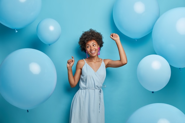 Felice donna dalla pelle scura gode di musica alla festa, balla spensierata, si diverte e si muove al ritmo di una canzone allegra, vestita con abiti festosi, isolata su un muro blu con palloncini decorati.