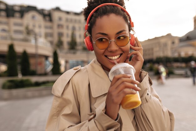 Felice donna dalla pelle scura in trench beige e occhiali beve succo d'arancia, ascolta musica in cuffia e sorride fuori