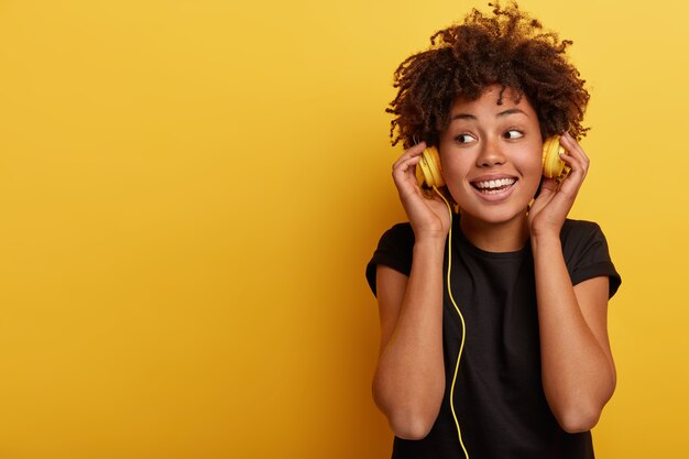 Felice donna dalla pelle scura indossa cuffie cablate, gode di buona musica, distoglie lo sguardo, ha un sorriso a trentadue denti