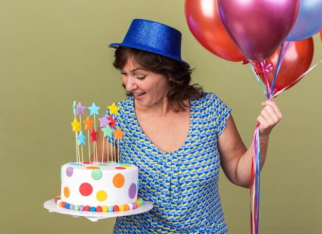 Felice donna di mezza età con cappello da festa con un mazzo di palloncini colorati con in mano una torta di compleanno guardandola sorridente che celebra la festa di compleanno in piedi sul muro verde green