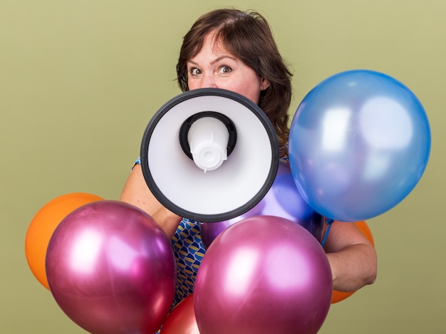 Felice donna di mezza età con un mucchio di palloncini colorati che gridano al megafono