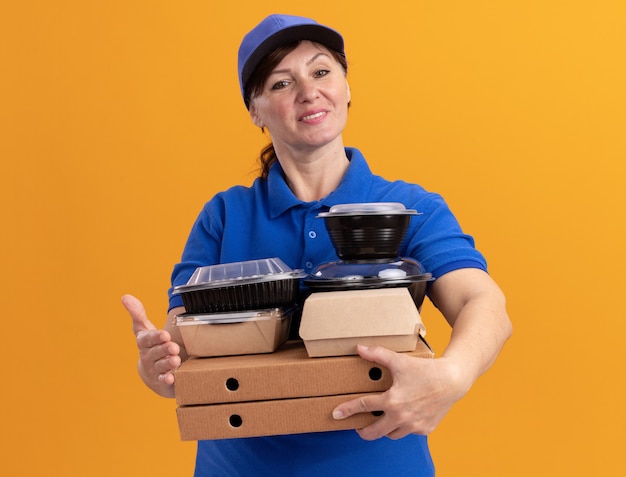 Felice donna di mezza età consegna in uniforme blu e cappuccio tenendo scatole per pizza e confezioni di cibo guardando davanti sorridente fiducioso in piedi sopra la parete arancione