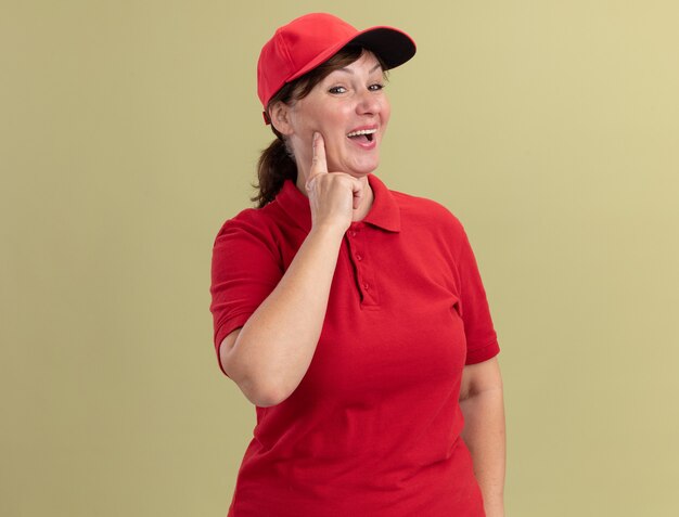 Felice donna di mezza età consegna in uniforme rossa e berretto guardando davanti sorridendo allegramente in piedi sopra la parete verde