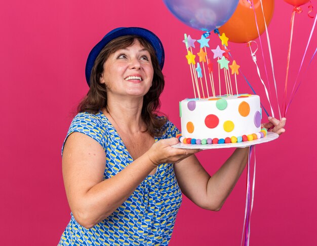 Felice donna di mezza età in cappello da festa con palloncini colorati che tengono la torta di compleanno alzando lo sguardo con un sorriso sul viso che celebra la festa di compleanno in piedi sul muro rosa