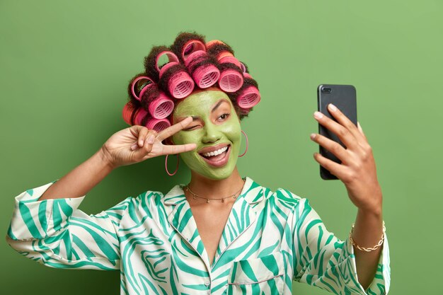 Felice donna divertente fa forme selfie segno di vittoria alla fotocamera dello smartphone sorride ampiamente gode di trattamenti per il visoapplica bigodini vestiti con abiti domestici casuali isolati sul muro verde