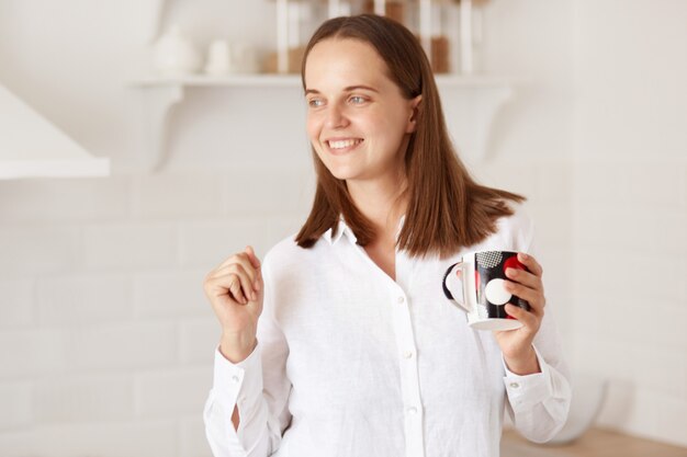 Felice donna eccitata che si sveglia presto la mattina, in piedi con una tazza di caffè o tè in mano e balla, di buon umore, in posa con set da cucina sullo sfondo.