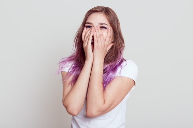 Felice donna elegante positiva che indossa la maglietta casual bianca con capelli lilla che ride felicemente, che copre il viso con le palme, esprimendo gioia, in posa isolato sopra il muro grigio