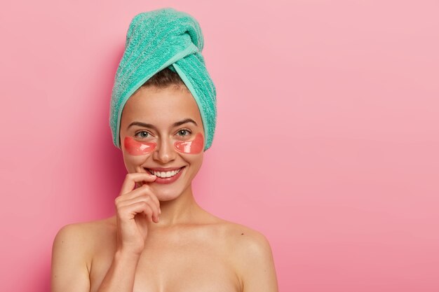 Felice donna europea si prende cura della pelle delicata intorno agli occhi, applica patch di collagene, indossa un trucco minimo, asciugamano da bagno avvolto sulla testa, sta nuda su sfondo rosa.