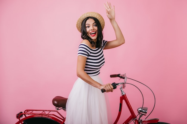 Felice donna in cappello di paglia in posa sulla bicicletta con un sorriso sorpreso. Tiro al coperto di attraente ragazza bruna che esprime emozioni felici.