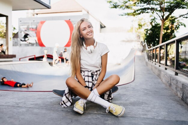 Felice donna magra seduta sul longboard dopo l'allenamento. Ritratto all'aperto del modello femminile abbastanza biondo che posa nello skate park nel fine settimana.