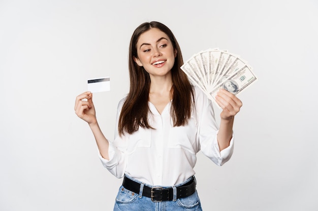 Felice donna moderna in possesso di denaro e carta di credito in mano, sorridente soddisfatto, in piedi su sfondo bianco. Copia spazio