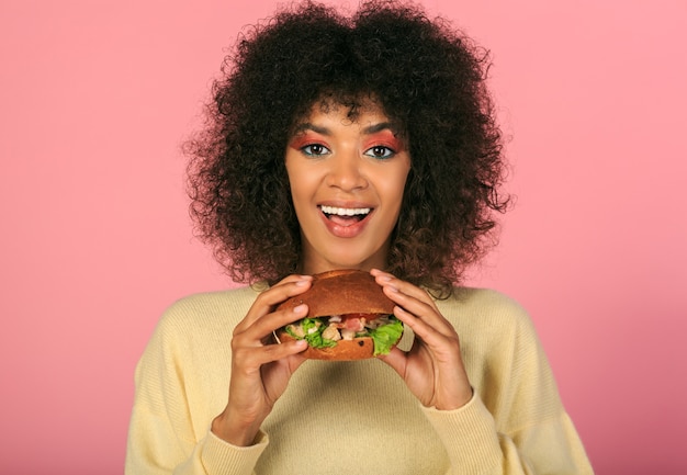 felice donna nera con i capelli ondulati mangiare gustosi cheeseburger sul rosa.