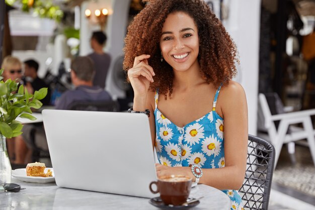 Felice donna professionista felice con i capelli ricci e la pelle scura, indossa una camicetta stampata a fiori blu, lavora su un computer portatile portatile