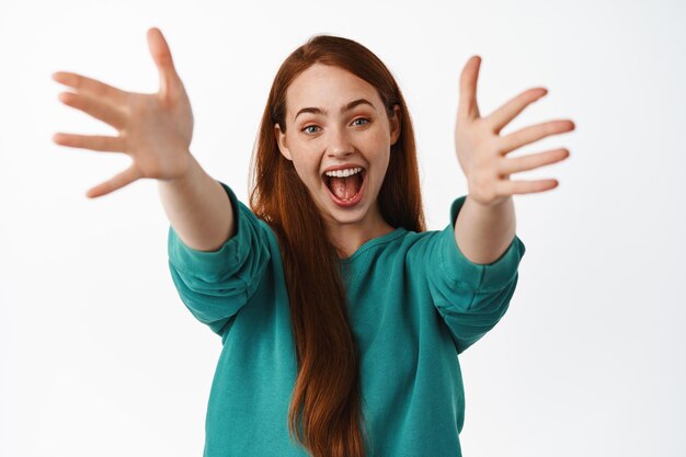 Felice donna rossa allunga le mani per abbracciare, raggiungendo abbracci caldi e amichevoli, sorridendo allegro, in piedi su sfondo bianco