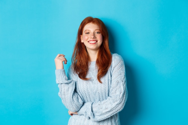 Felice donna rossa in maglione, che guarda con piacere la macchina fotografica e sorride, in piedi su sfondo blu.