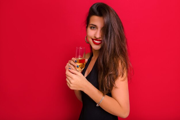 Felice donna sorridente su sfondo rosso che celebra il nuovo anno, indossando abiti di lusso e tenendo champagne.