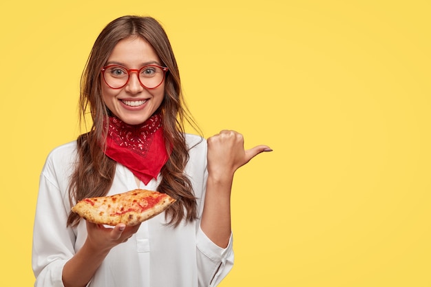 Felice donna sorridente tiene una gustosa pizza, indica con il pollice da parte come mostra il luogo in cui l'ha comprata, fa pubblicità alla pizzeria, indossa una bandana rossa e una camicia bianca, isolata su un muro giallo.