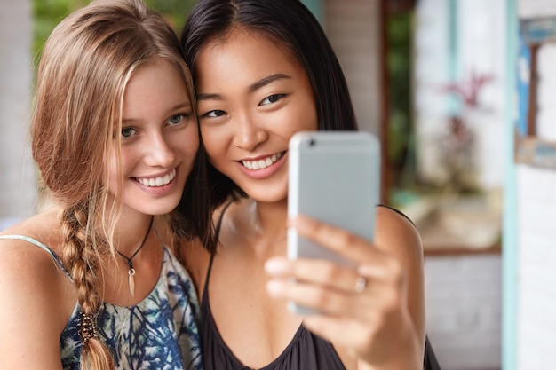 Felice due donne di razza mista sorridono e posano davanti alla telecamera, usano il cellulare moderno per fare foto, abbracciano e ridono allegramente