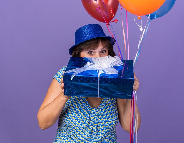 Felice e allegra donna di mezza età con cappello da festa che tiene in mano un mazzo di palloncini colorati e presenta un aspetto incuriosito